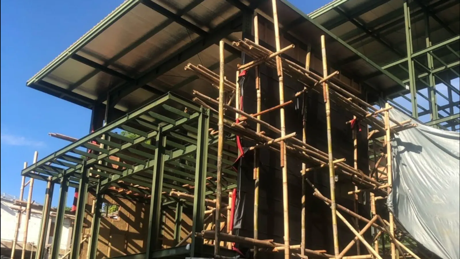 Green steel columns and roof frame of a two-level building surrounded by bamboo scaffolding